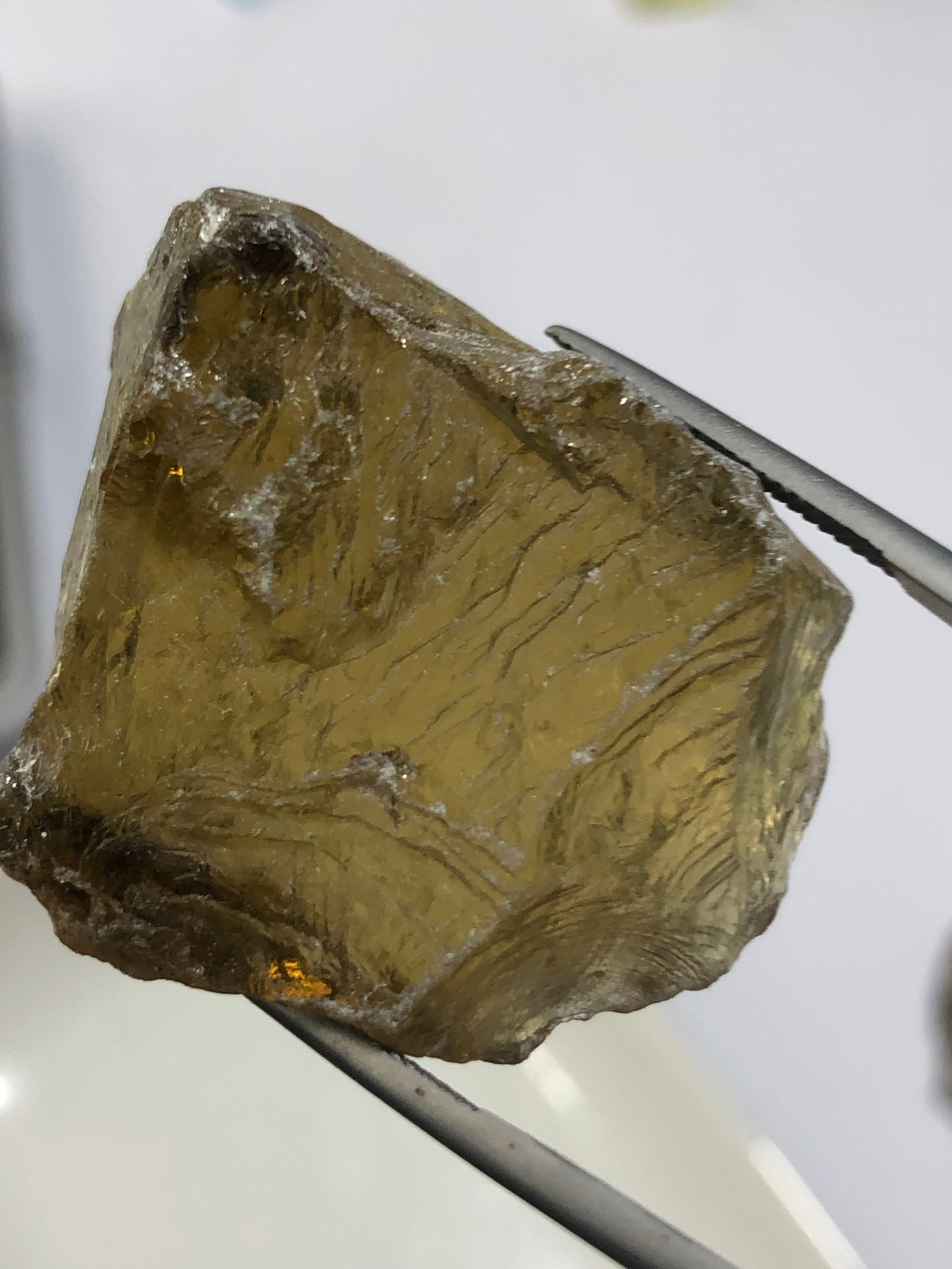 A photograph of a raw smoky citrine stone held by tweezers, displaying its rough, uncut form with a smoky color.